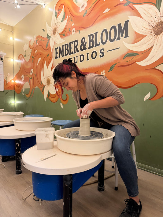 A woman with red hair in a brown cardigan and blue jeans sits at a pottery wheel, shaping a clay vase. The setting is a pottery studio with a green wall decorated with orange and white floral artwork that reads 'EMBER & BLOOM STUDIOS'.