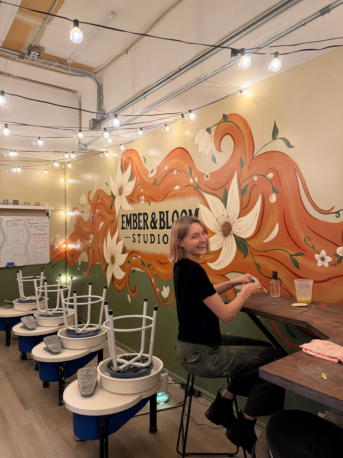 A woman sits at a wooden counter inside a creative studio with a colorful mural on the wall reading 'EMBER & BLOOM STUDIO'. The room features pottery wheels stacked with white stools, hanging string lights, and an industrial ceiling.