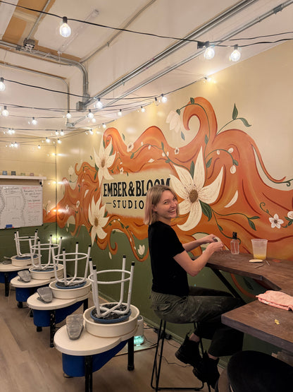 A woman sits at a wooden counter inside a creative studio with a colorful mural on the wall reading 'EMBER & BLOOM STUDIO'. The room features pottery wheels stacked with white stools, hanging string lights, and an industrial ceiling.